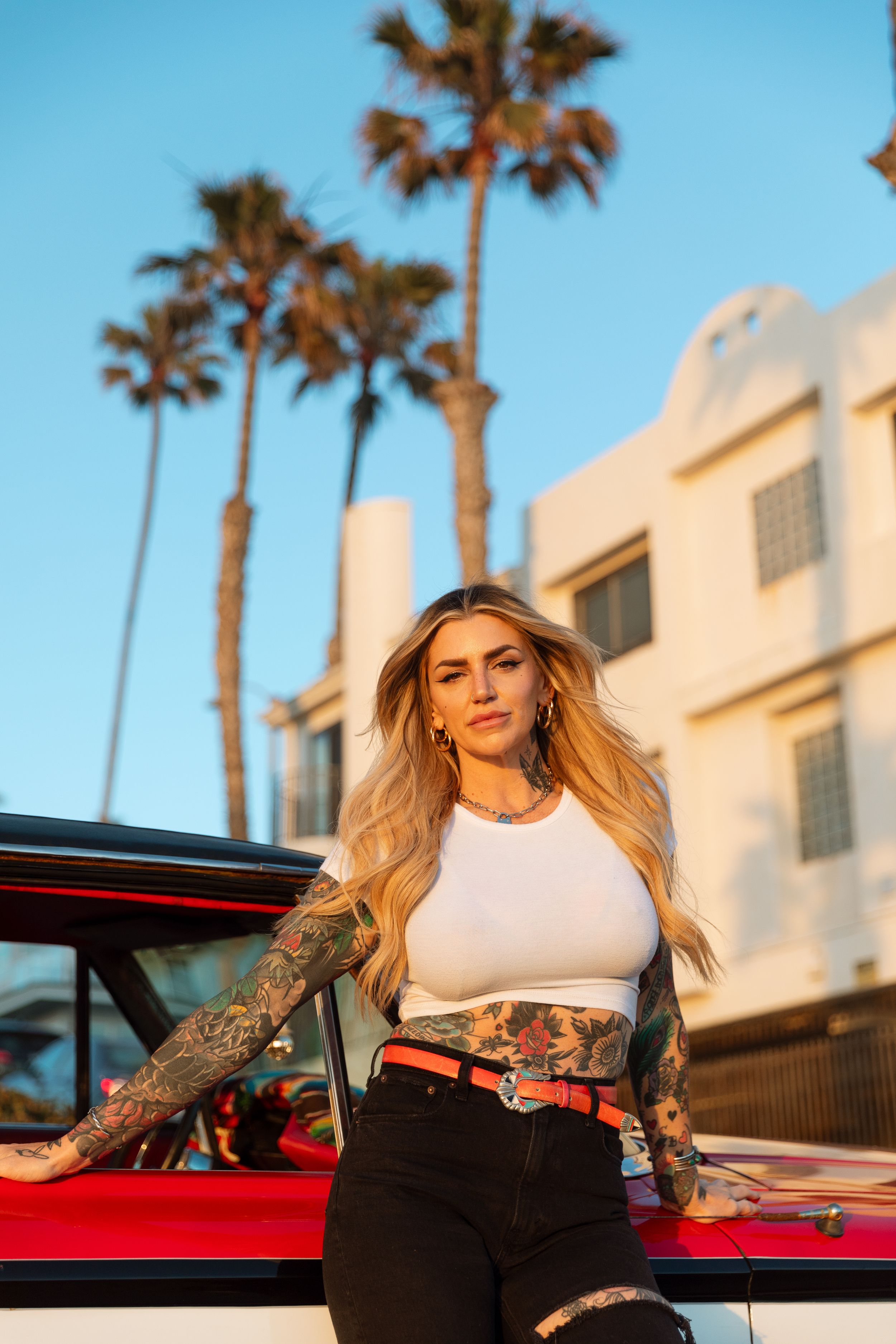 A woman in a white crop top and black pants poses against a red car with palm trees and art deco building in Miami.