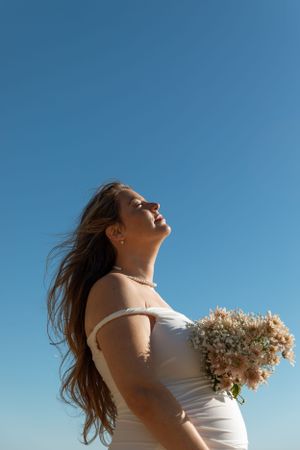 A pregnant woman in a white dress holds a dried flower bouquet against a bright blue sky on a sunny day.