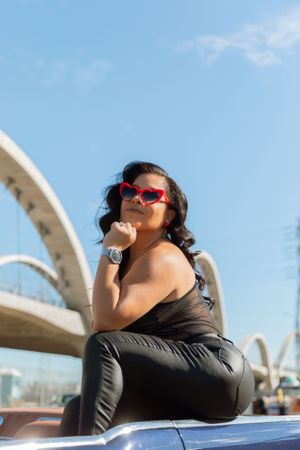 Person in pink sunglasses and black outfit poses on car roof with curved architectural structure in background.