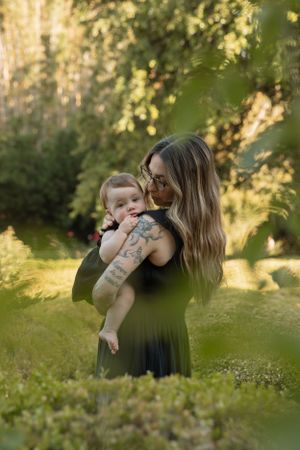 Mother in black dress holds baby while standing in sunlit garden with lush greenery.