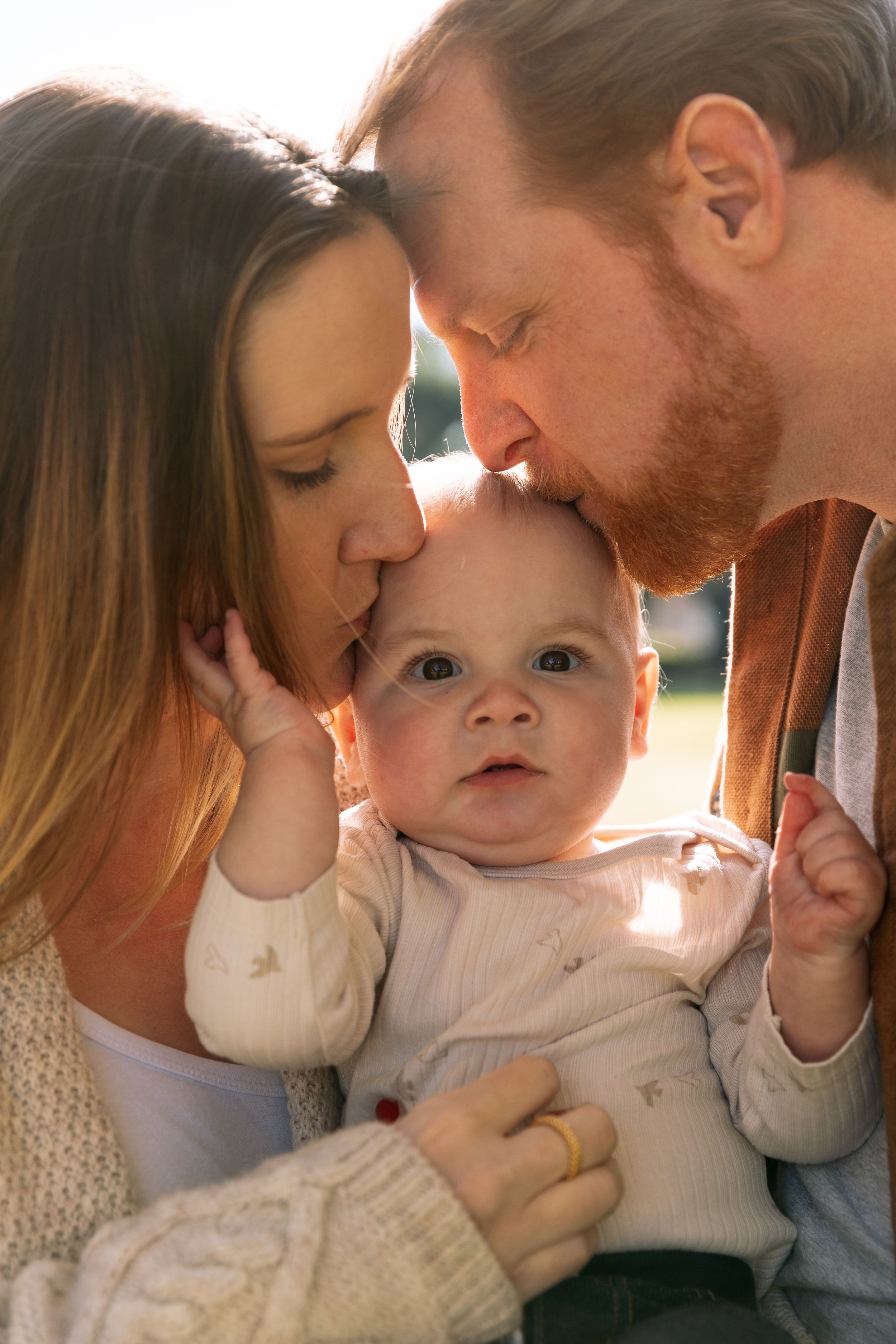 A small infant is held and kissed by two adults in soft, warm sunlight.