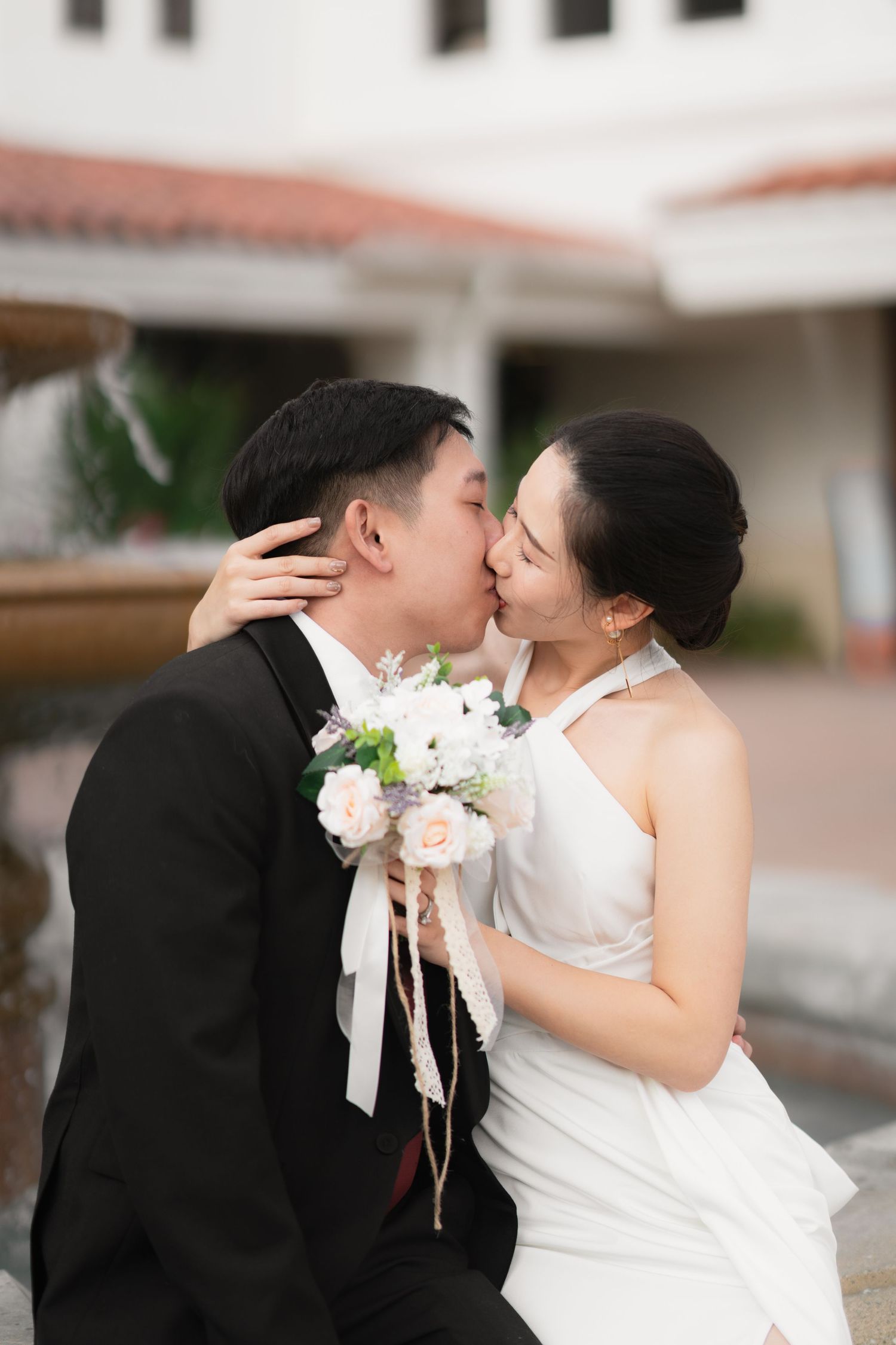 Asian bride and groom sharing a kiss outdoors near a fountain, bride holding floral bouquet.