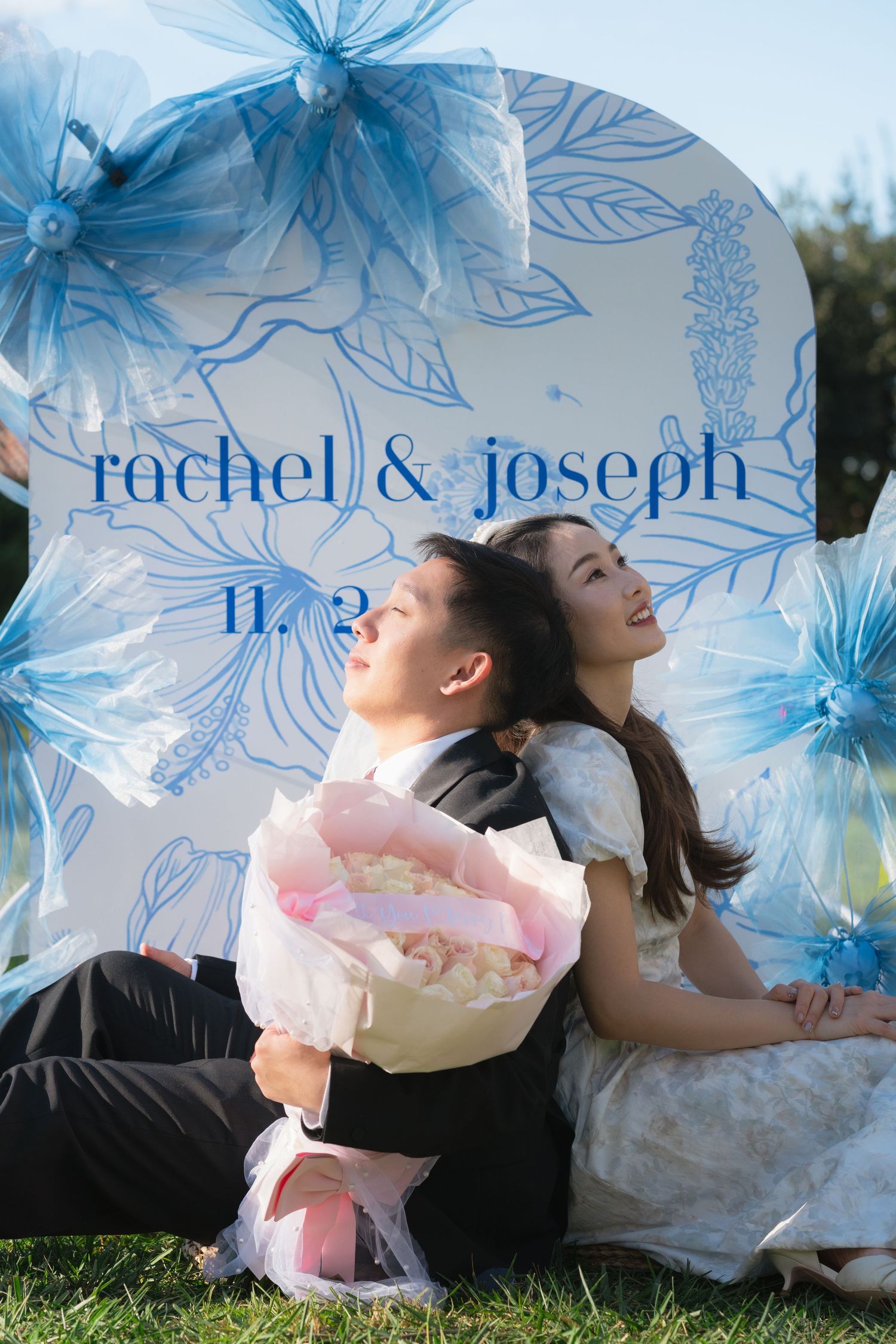 Rachel and Joseph sit back-to-back on grass before a floral wedding sign, groom holding a pink bouquet.