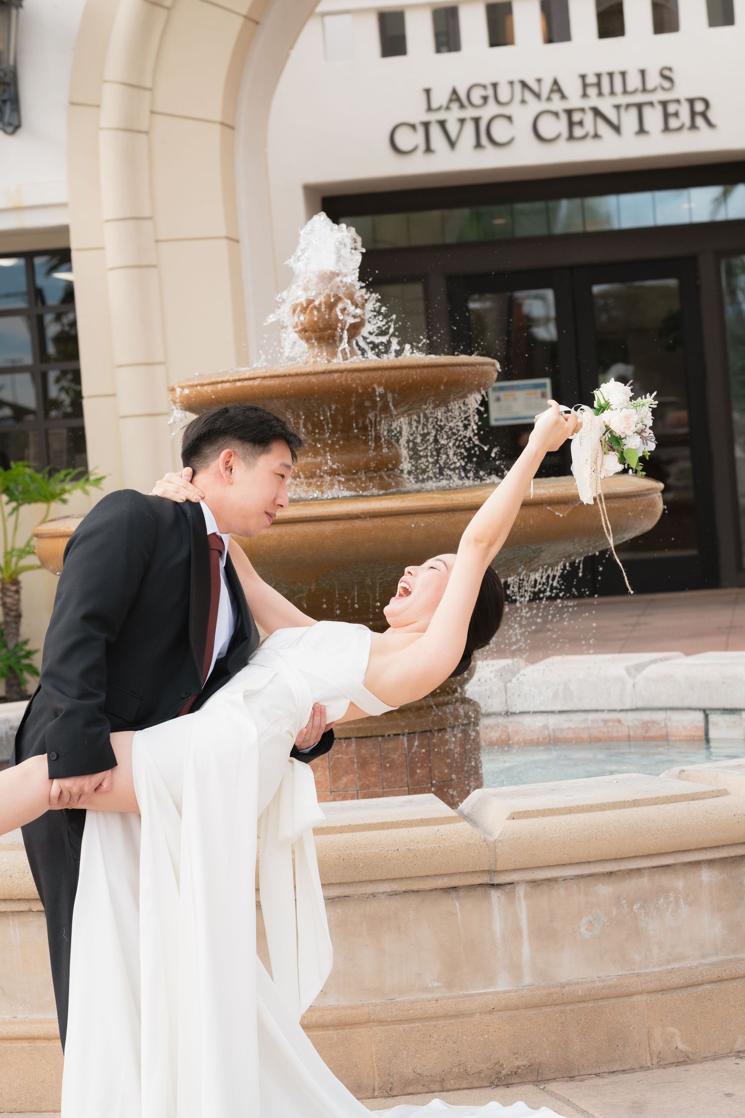 Asian couple in wedding attire dancing by a fountain at Laguna Hills Civic Center.