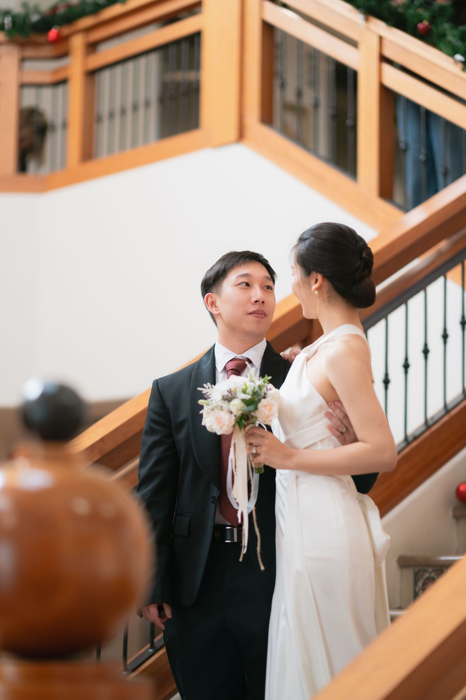 Asian bride and groom standing on staircase, exchanging glances while bride holds white bouquet.