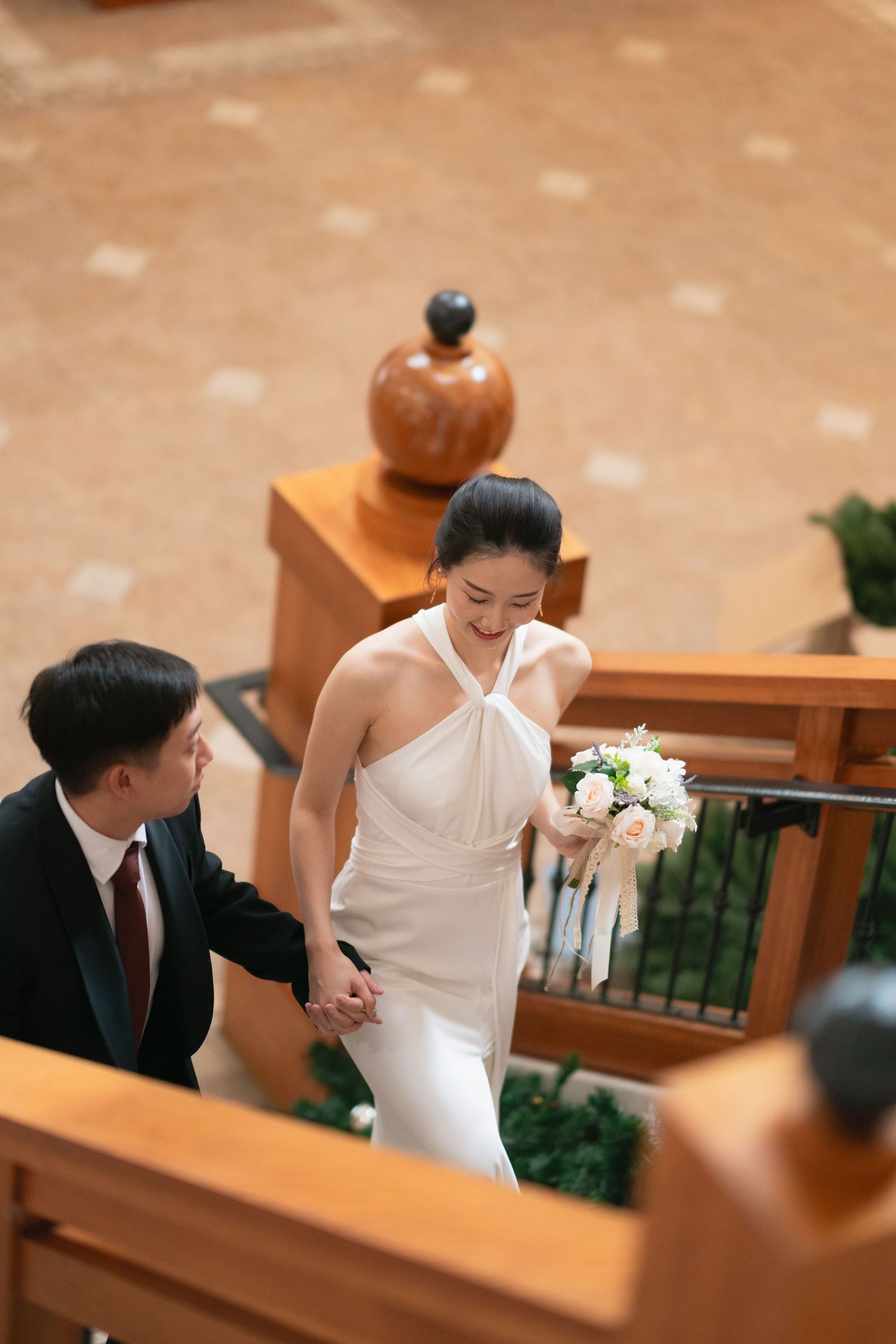 Aerial view of Asian bride and groom holding hands while ascending wooden staircase indoors.