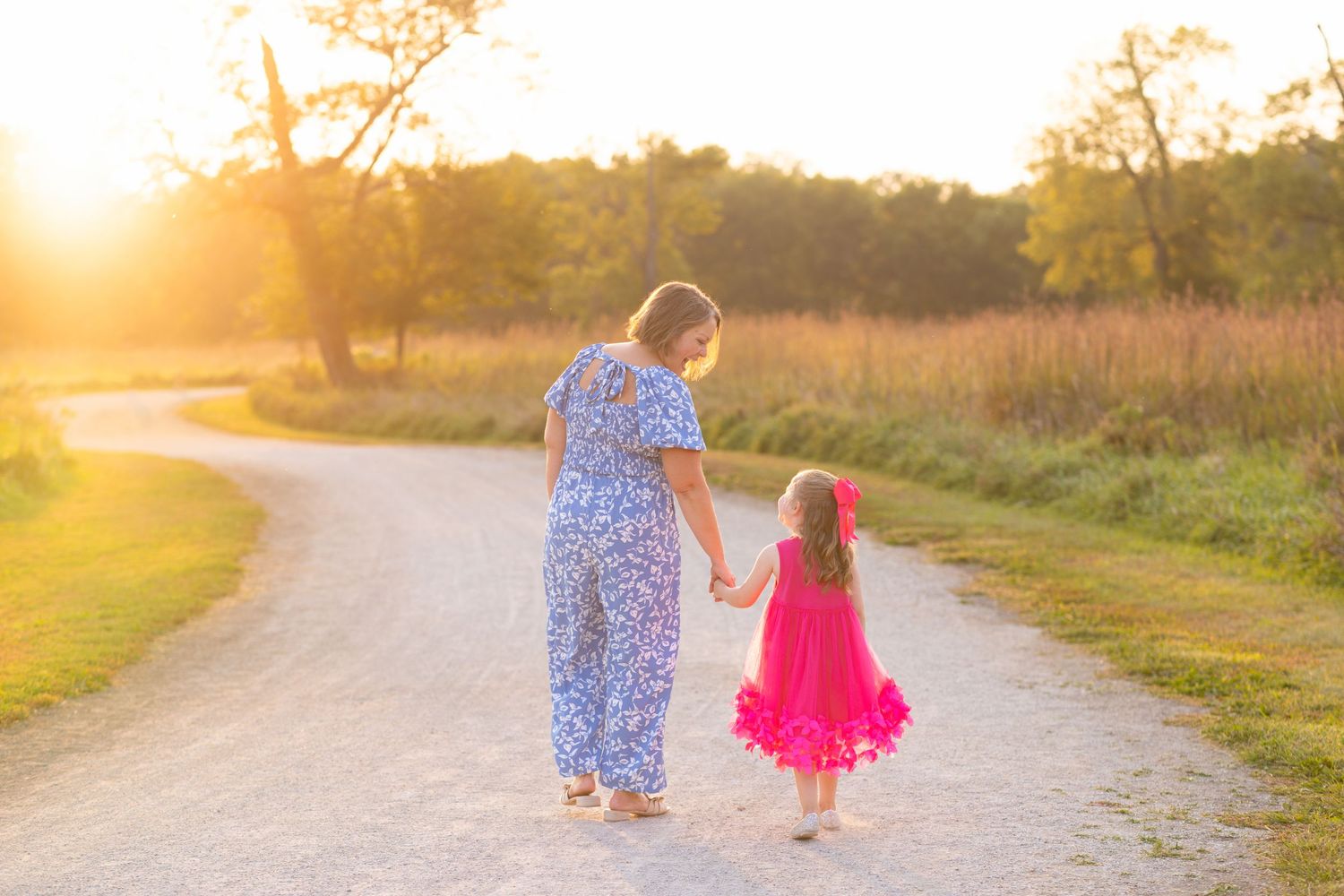 Family portrait of a mother in blue and a daughter in pink holding hands walking along a sunlit dirt path at golden hour.
