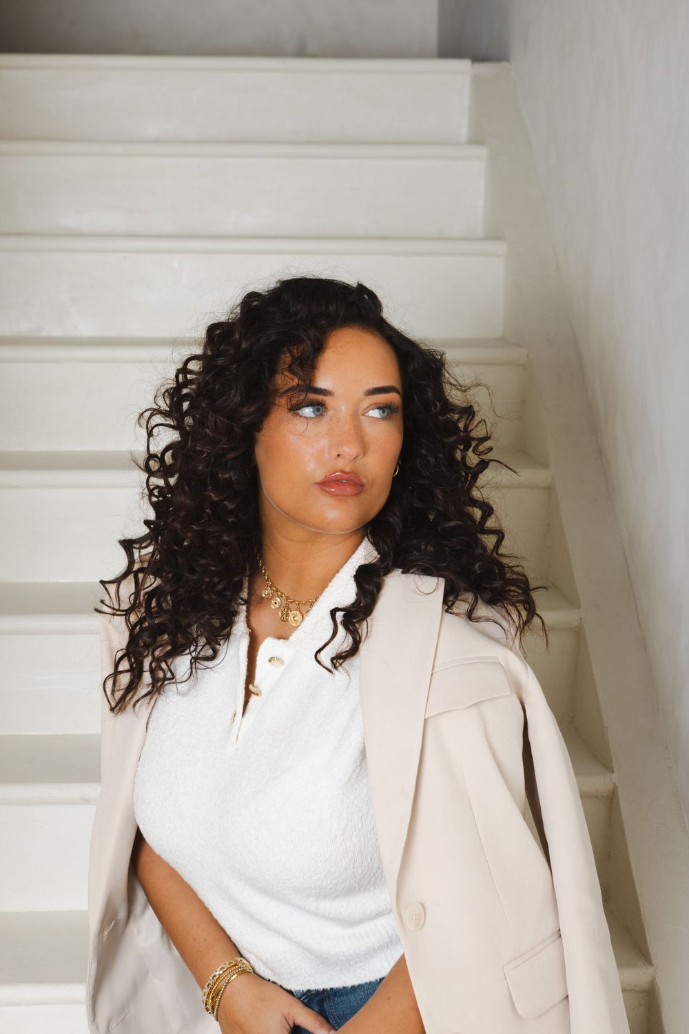 Model wearing white collared top and blazer poses elegantly against minimalist background.