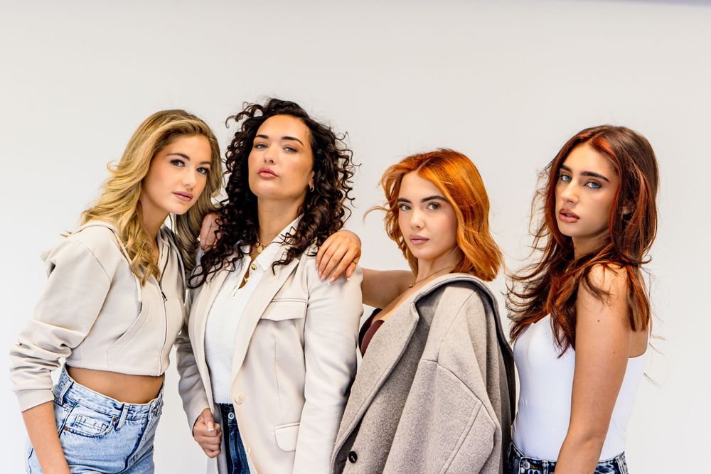 Four women with different hair colors pose together wearing casual outfits against a white background.
