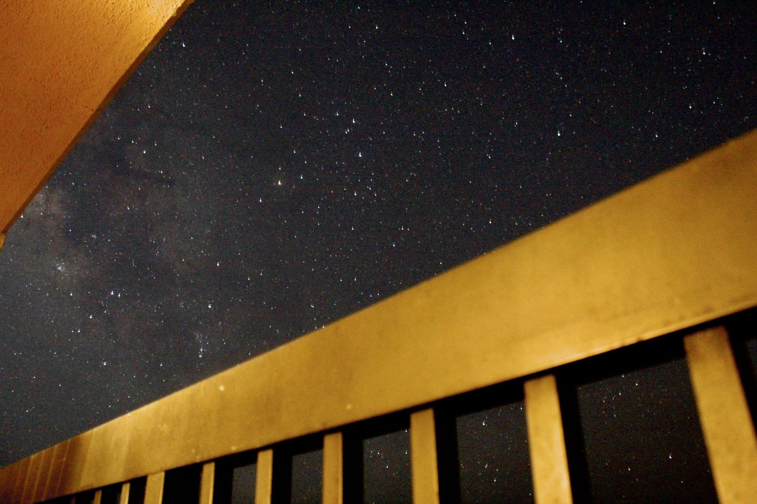 Night sky filled with stars viewed from beneath a golden-lit building balcony railing.