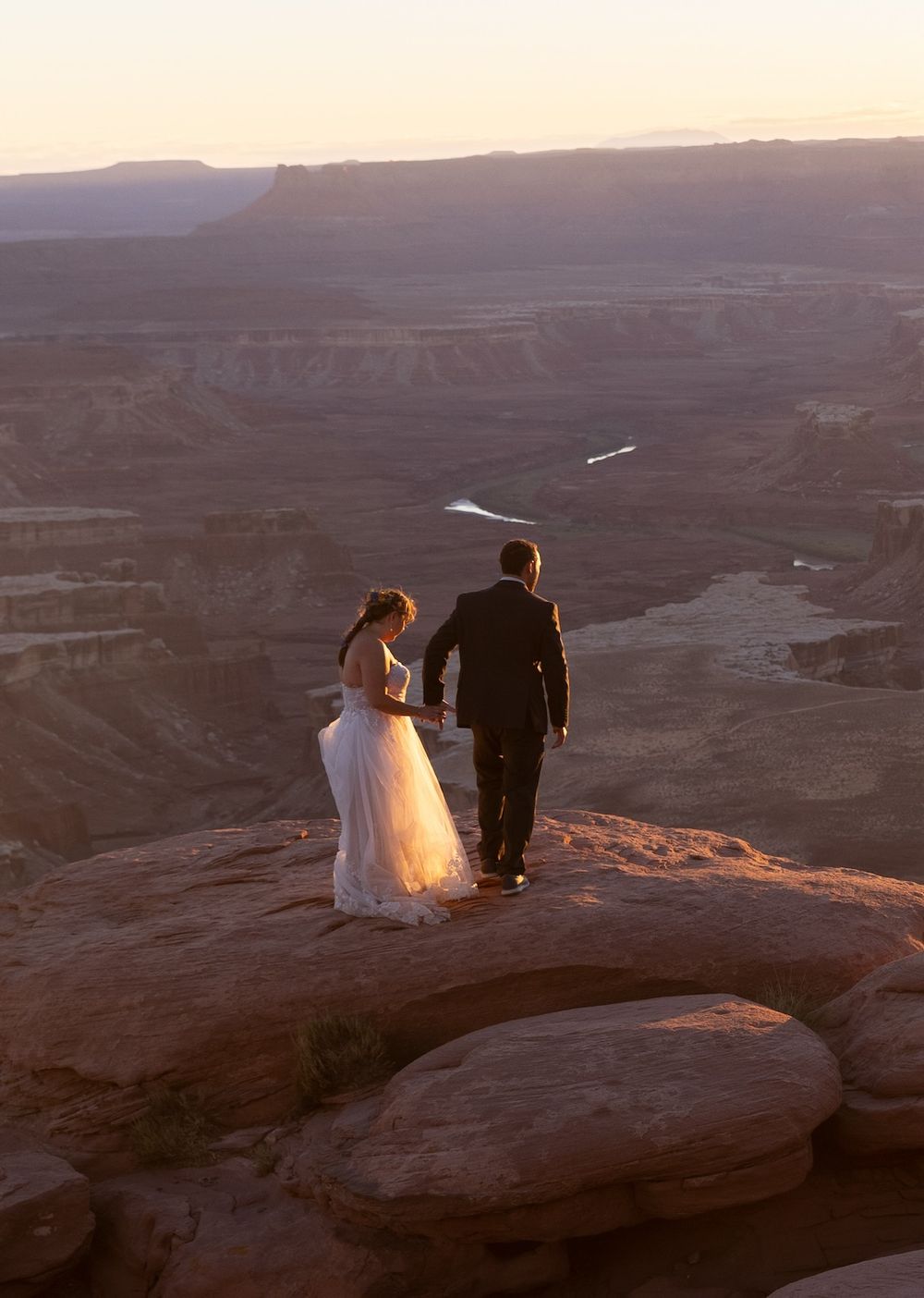 A couple sits at the edge of a cliff overlooking the vast desert canyons at sunset in Dead Horse Point State Park, Utah.
