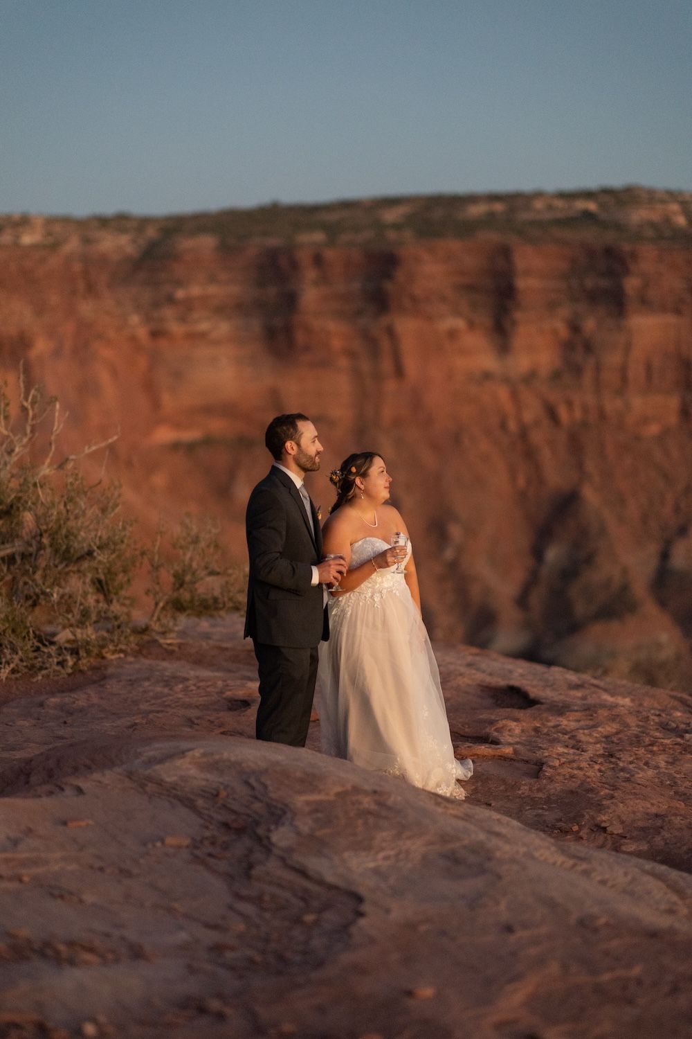 A couple shares a romantic moment at sunset on red rock cliffs overlooking a desert canyon landscape.