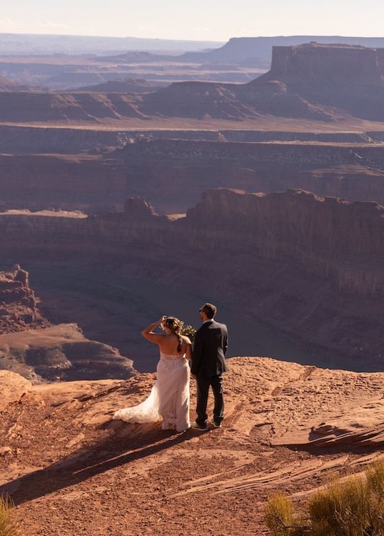 Two figures stand at the edge of a scenic desert canyon overlook during golden hour with vast mesas in the background.