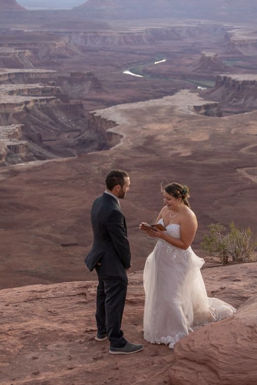 A couple in wedding attire sits on red rocks overlooking the vast canyonlands of Utah during their elopement ceremony.