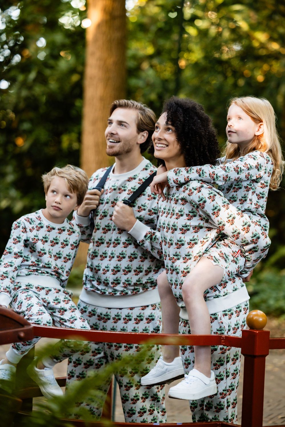 A family wearing matching patterned sweaters poses playfully on a wooden bench in a park setting at sunset.