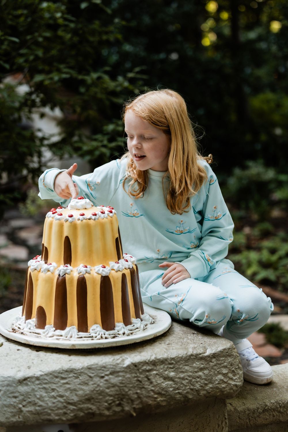 A golden bundt cake topped with whipped cream sits on a stone ledge in an outdoor garden setting.