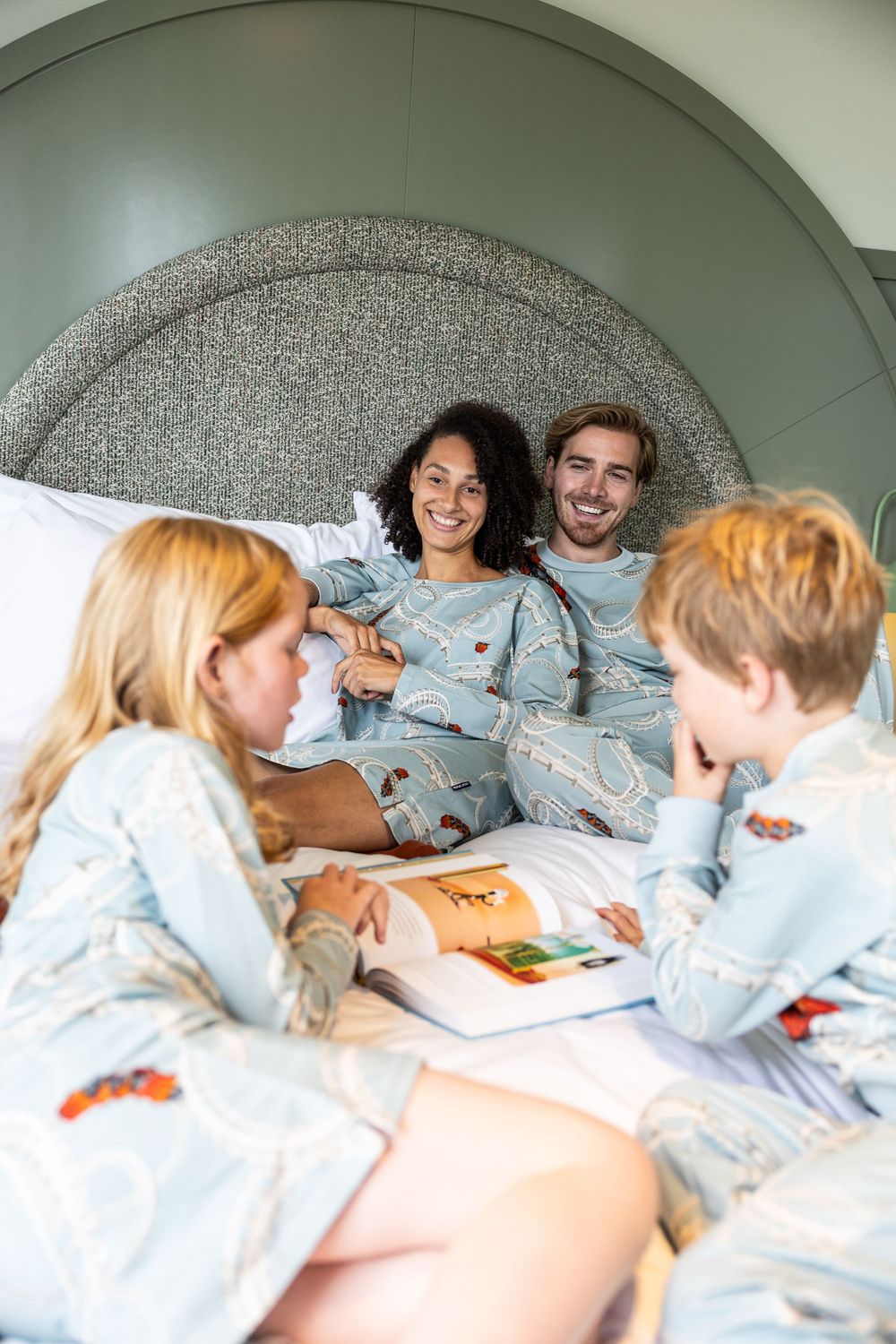 A family wearing matching light blue pajamas relaxes together on a large bed while reading books.