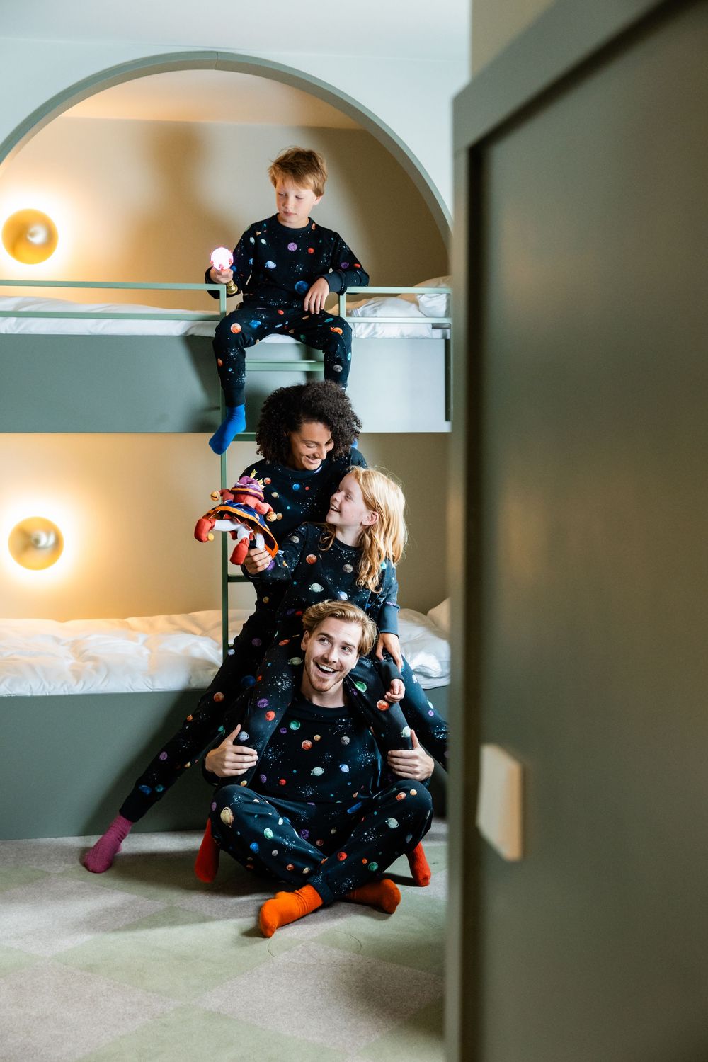 Four people wearing matching space-themed pajamas pose playfully on built-in bunk beds with warm lighting.