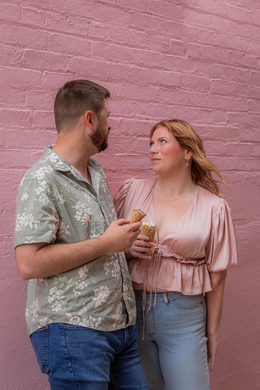 A couple in coordinating spring outfits share an ice cream cone while standing against a pink brick wall.