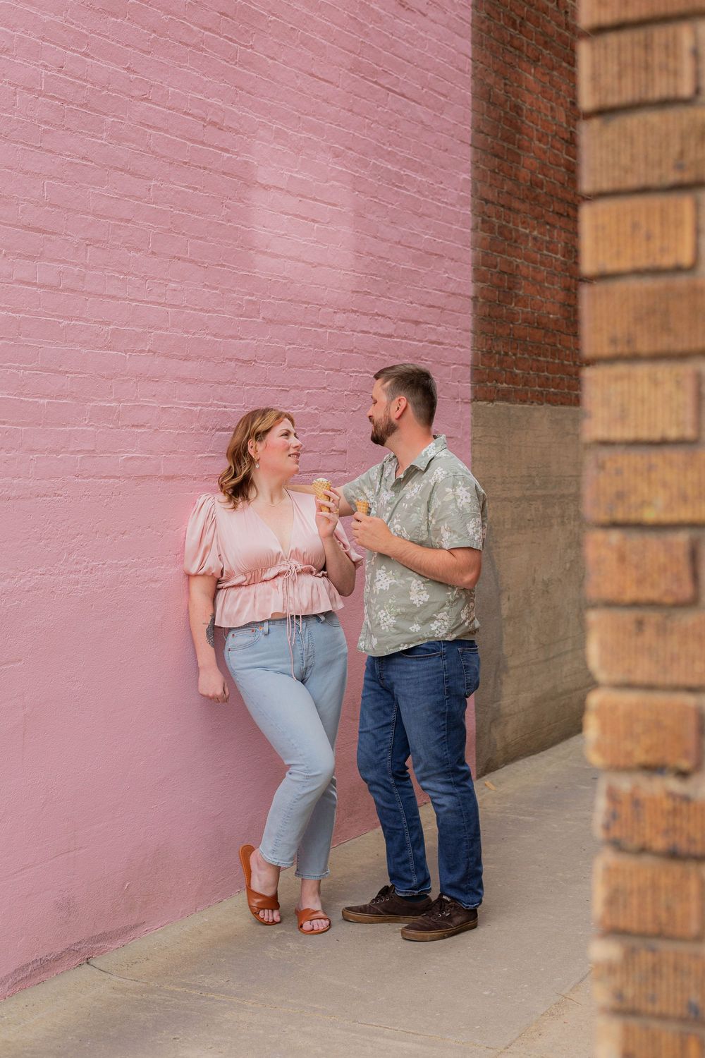 A couple enjoying ice cream while standing against a pink brick wall.