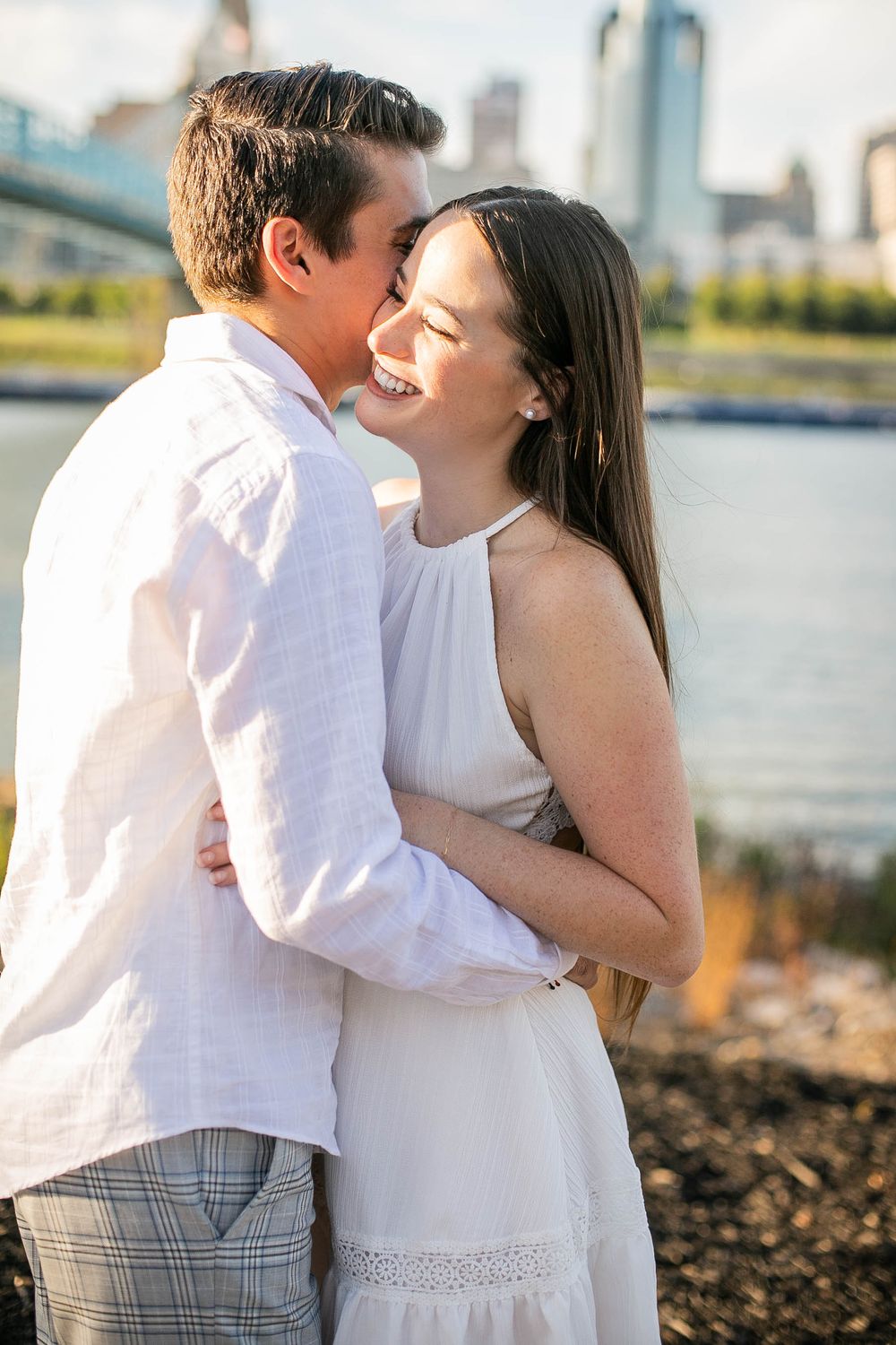 A romantic couple embraces outdoors by the waterfront at sunset, wearing white clothing with a city skyline in the background.