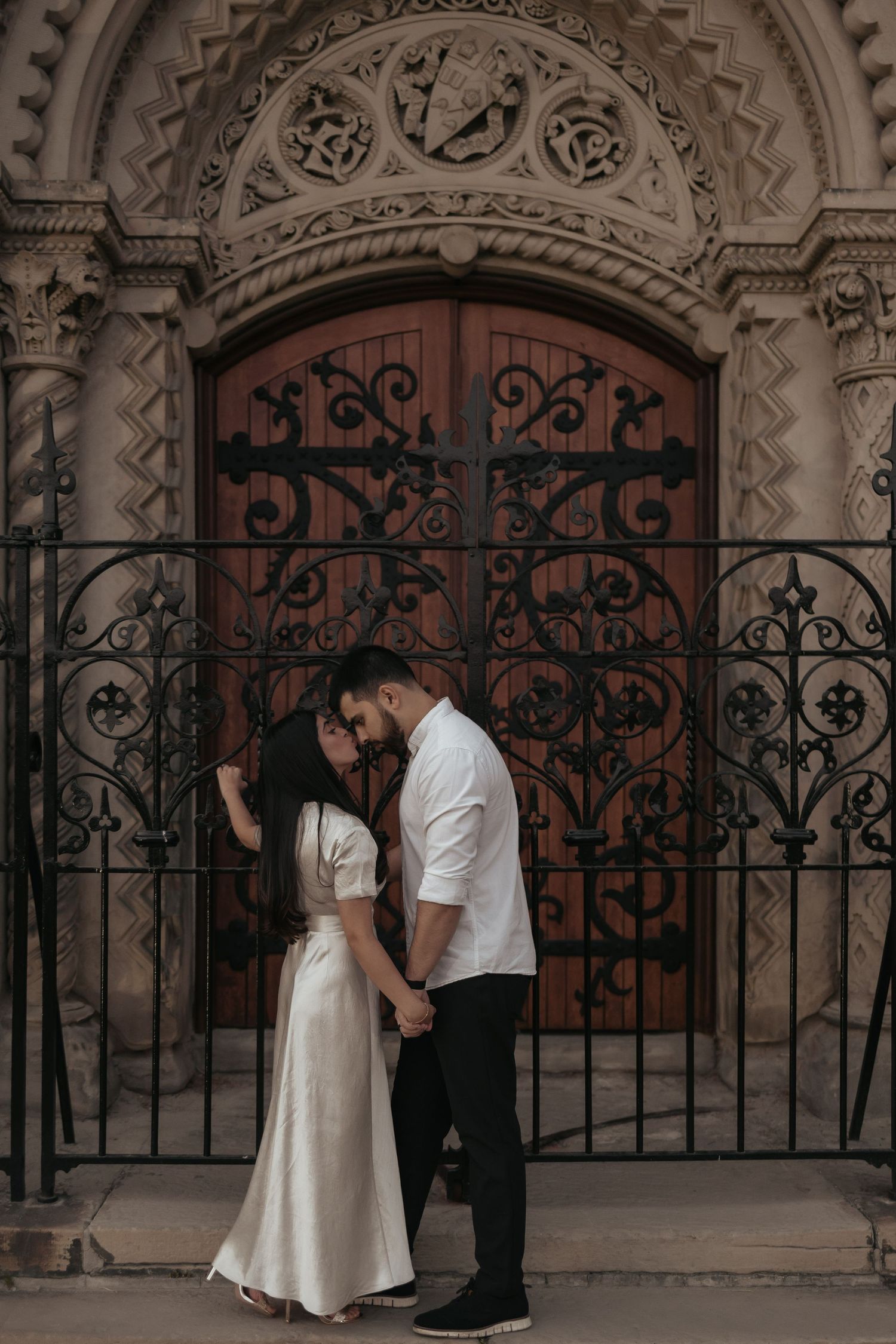 Romantic couple in formal attire pose against ornate vintage door with wrought iron details.
