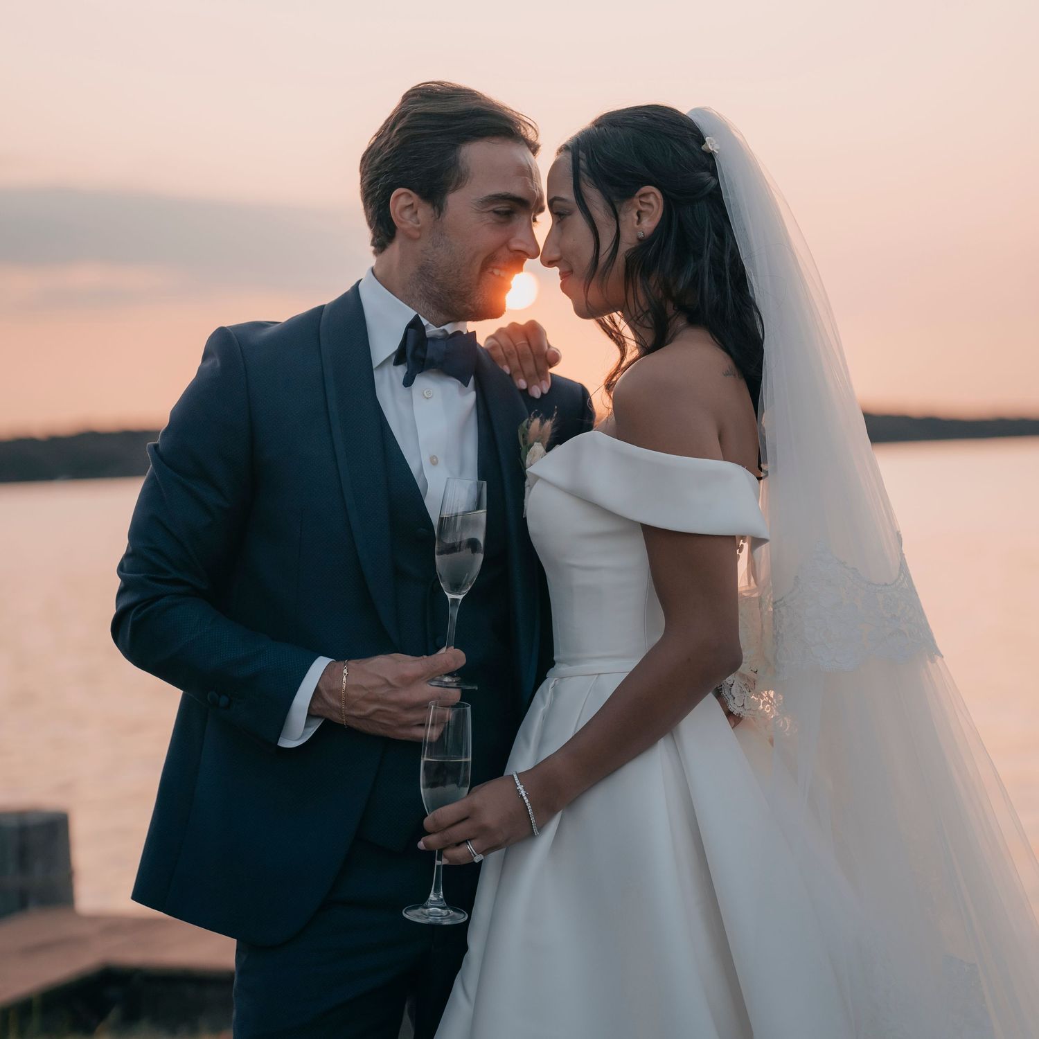 Romantic sunset portrait of newlyweds sharing champagne by waterfront.