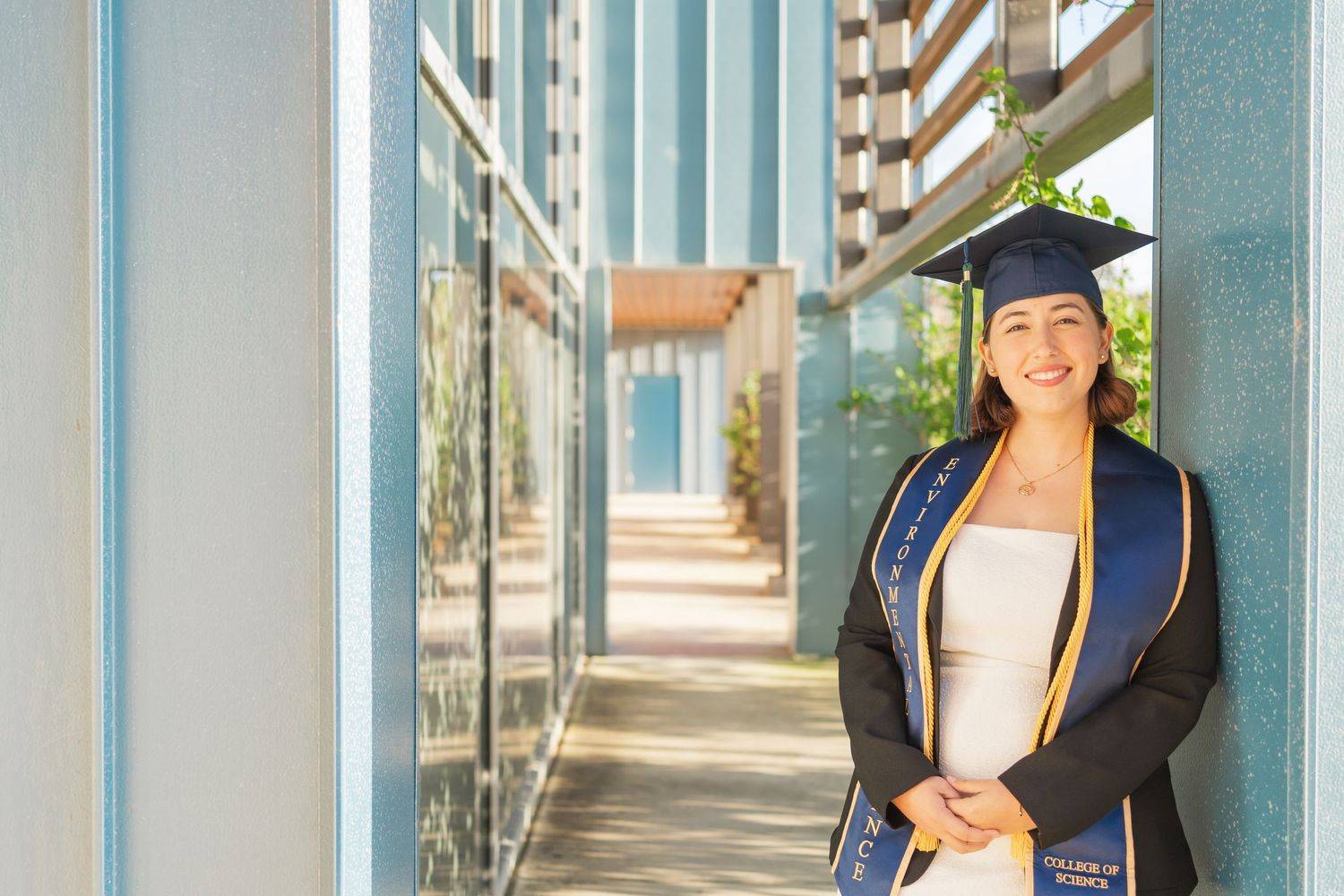 A graduate in cap and gown smiles while standing in a modern building hallway with glass windows and columns.