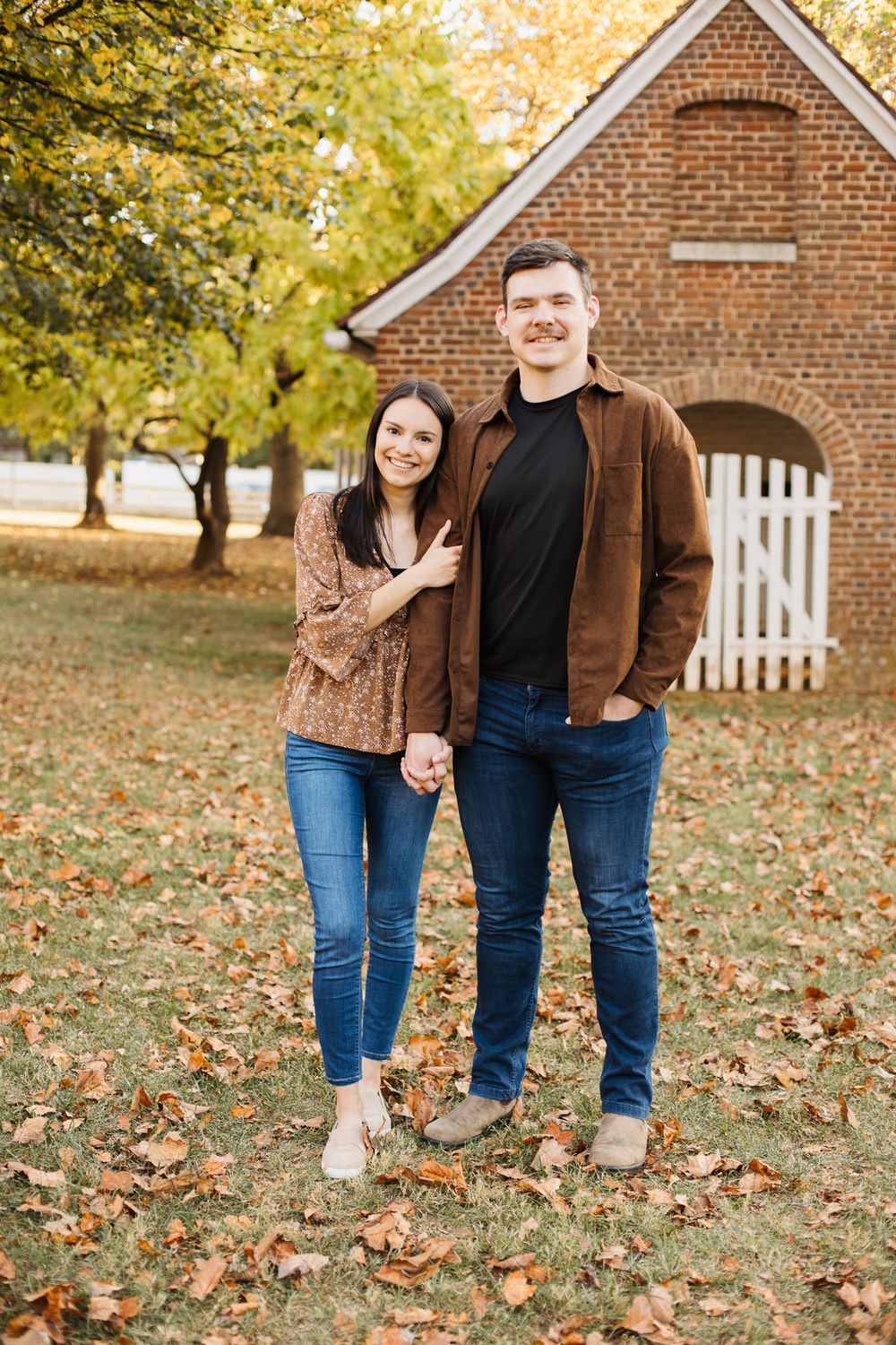 Couple in casual fall attire poses together on lawn with brick building and white fence in background.