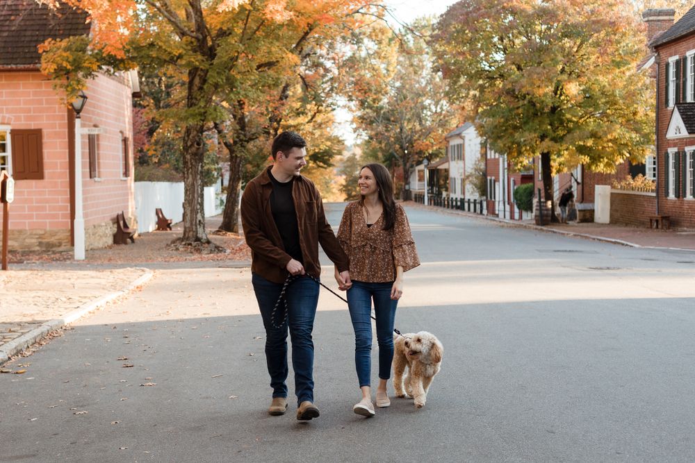 A couple and their golden retriever walk along a tree-lined path covered in autumn leaves.