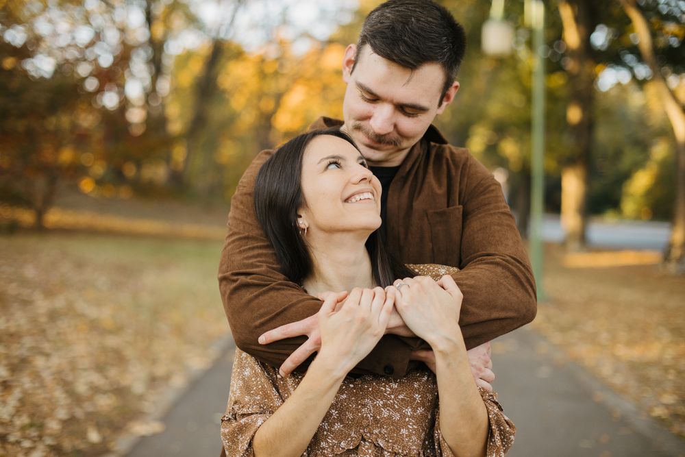 Two people share an intimate moment on a path with autumn trees in the background.