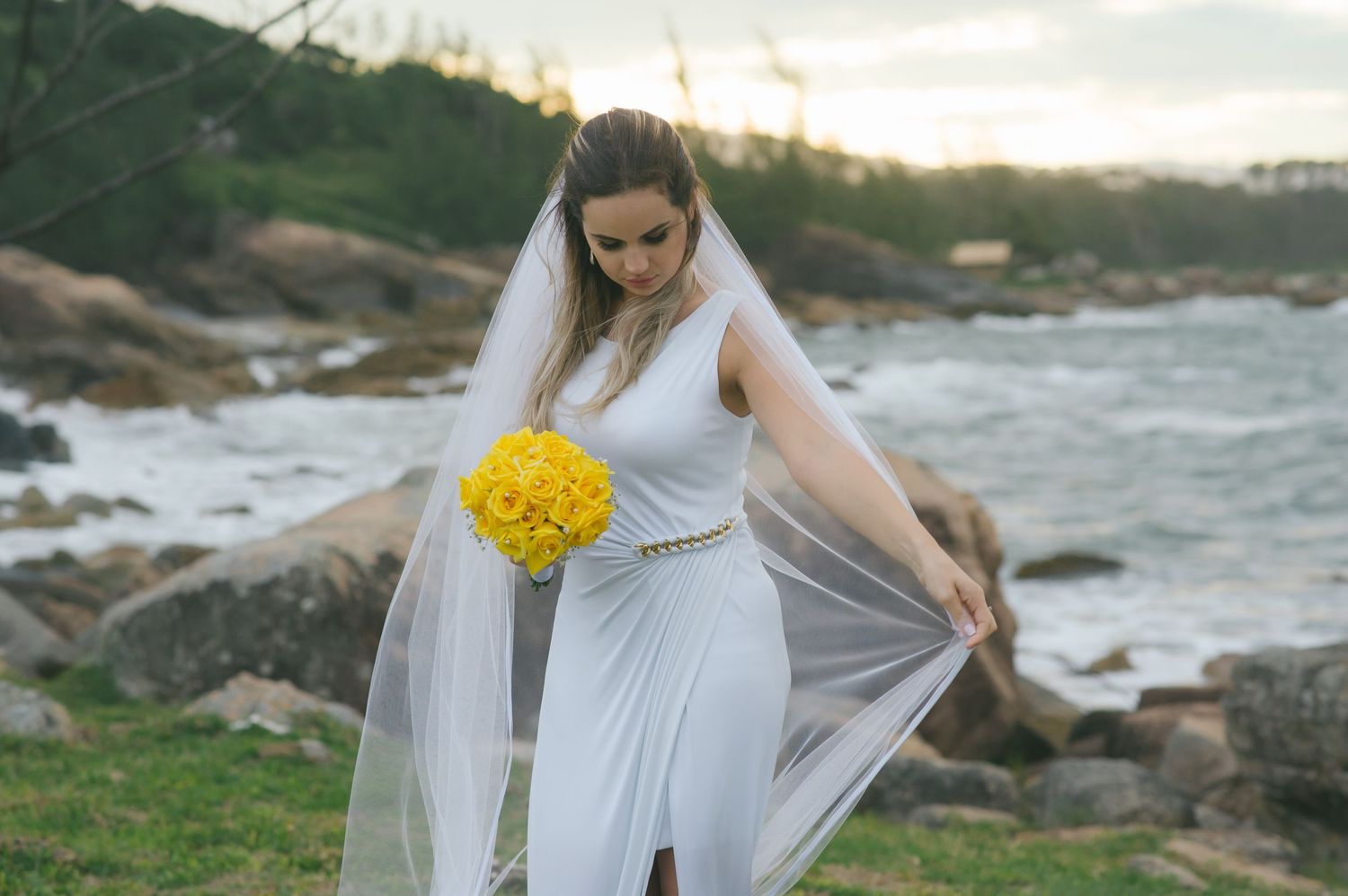 Elegant bride with yellow bouquet and flowing dress poses by rocky coastline.