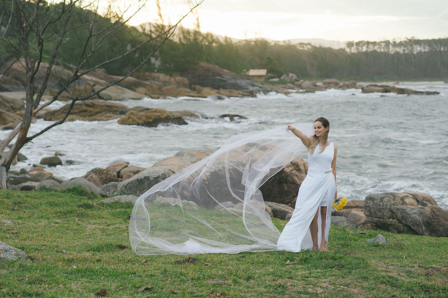 A flowing white wedding dress veil catches the wind against a rocky coastal backdrop at sunset.