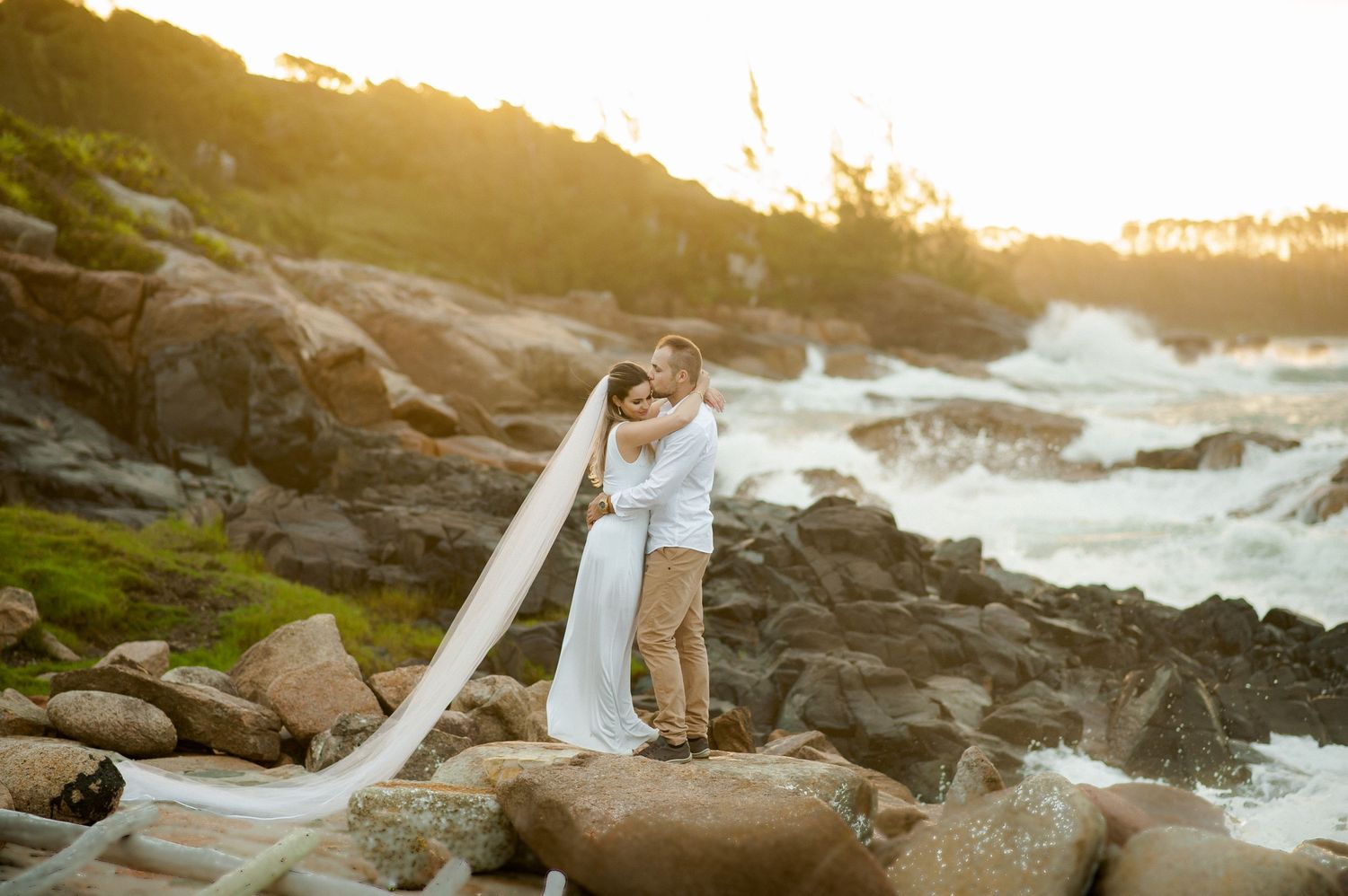 A wedding couple embraces on rocky coastal cliffs during golden hour.