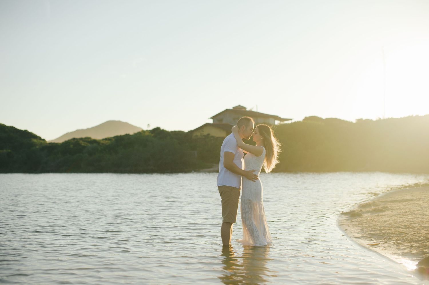 A couple stands together in shallow water at the edge of a lake during sunset.