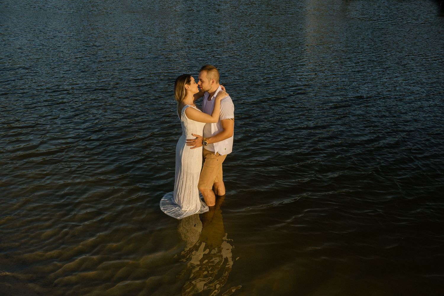 A couple embraces while standing in dark water during a sunset photoshoot.