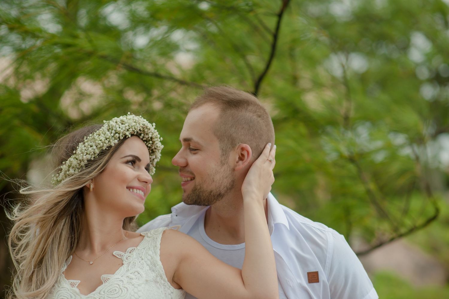 Couple in white attire sharing intimate moment under green foliage.