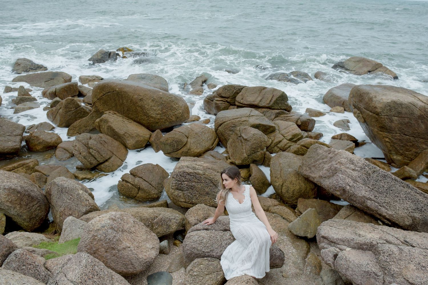 Woman in white dress sits alone on coastal rocks with crashing waves.