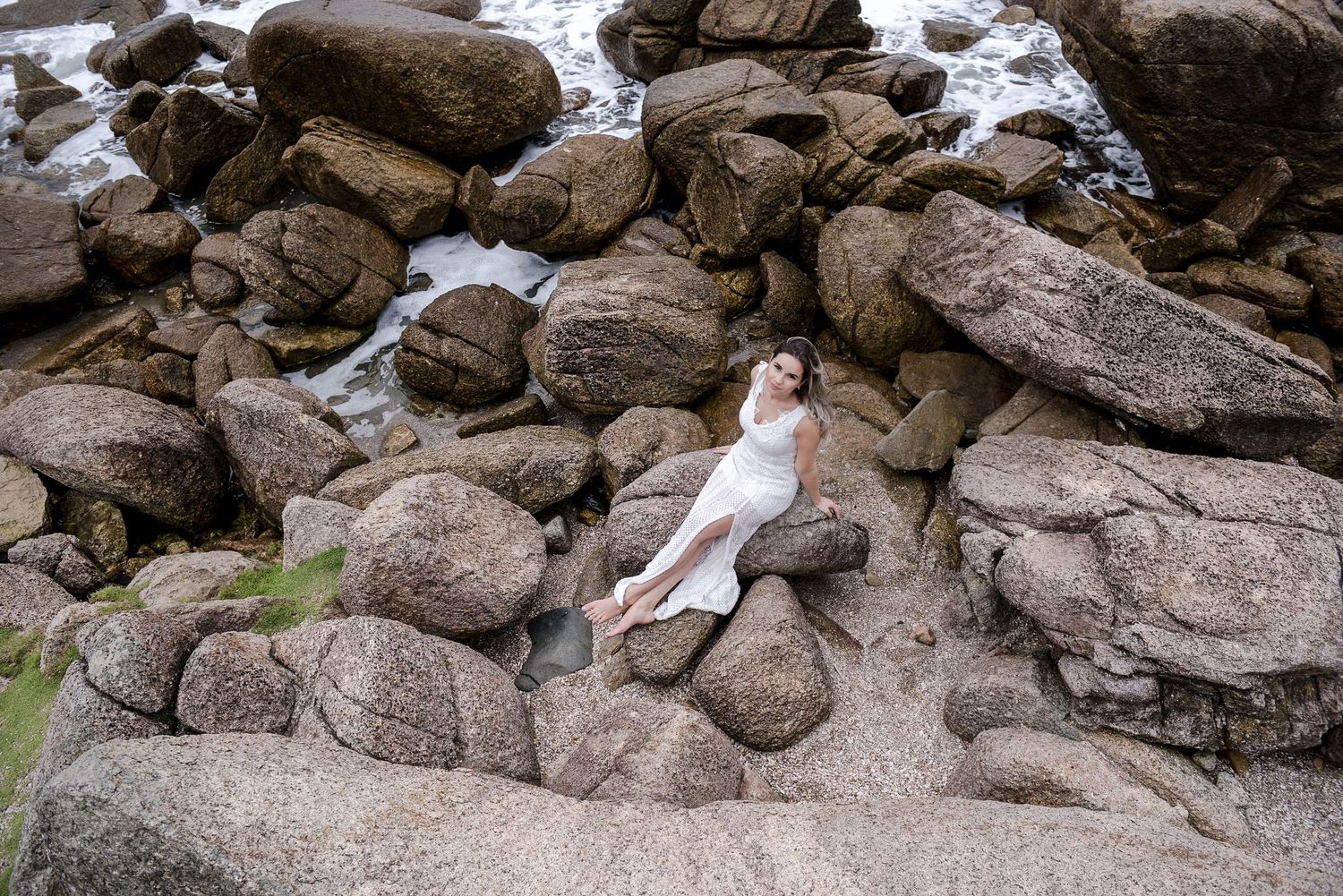 Woman in white dress reclines on rocky coastal formation.