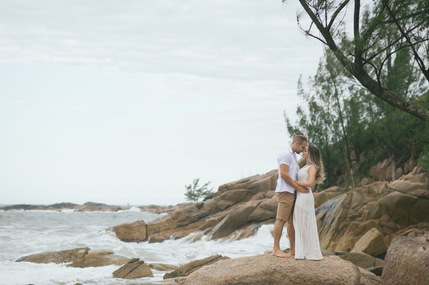 Couple in white stands on rocky coastal outcrop against ocean backdrop.