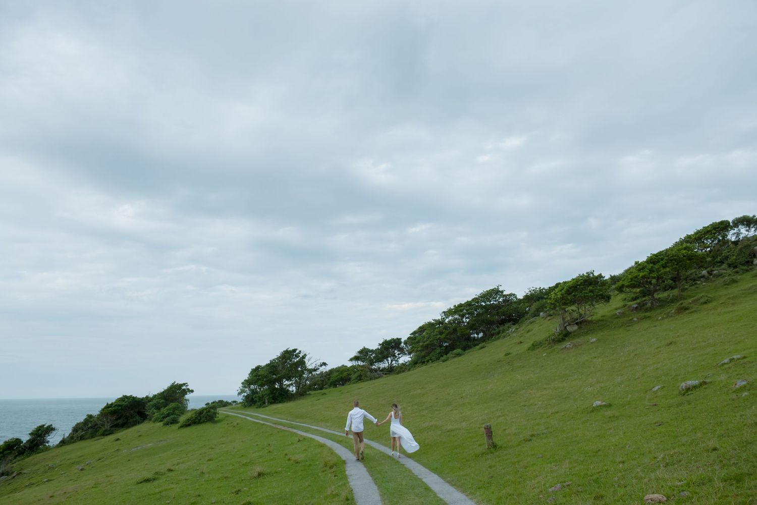 Two figures walk along winding coastal path on green hillside.