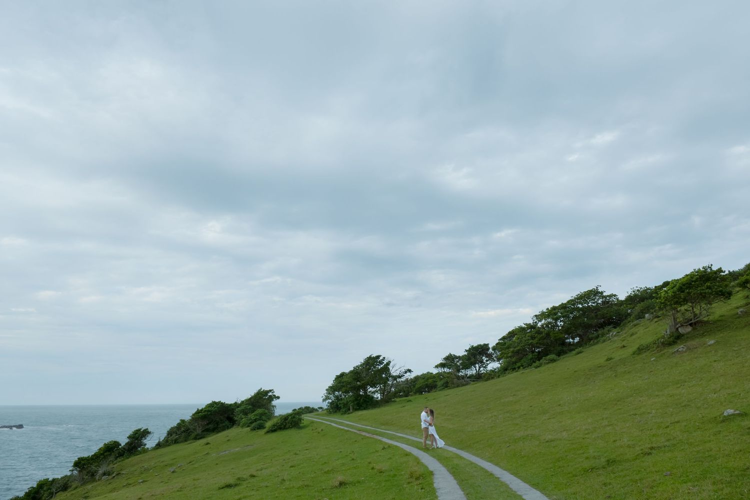 Person in flowing dress walks along curved seaside path on green hill.
