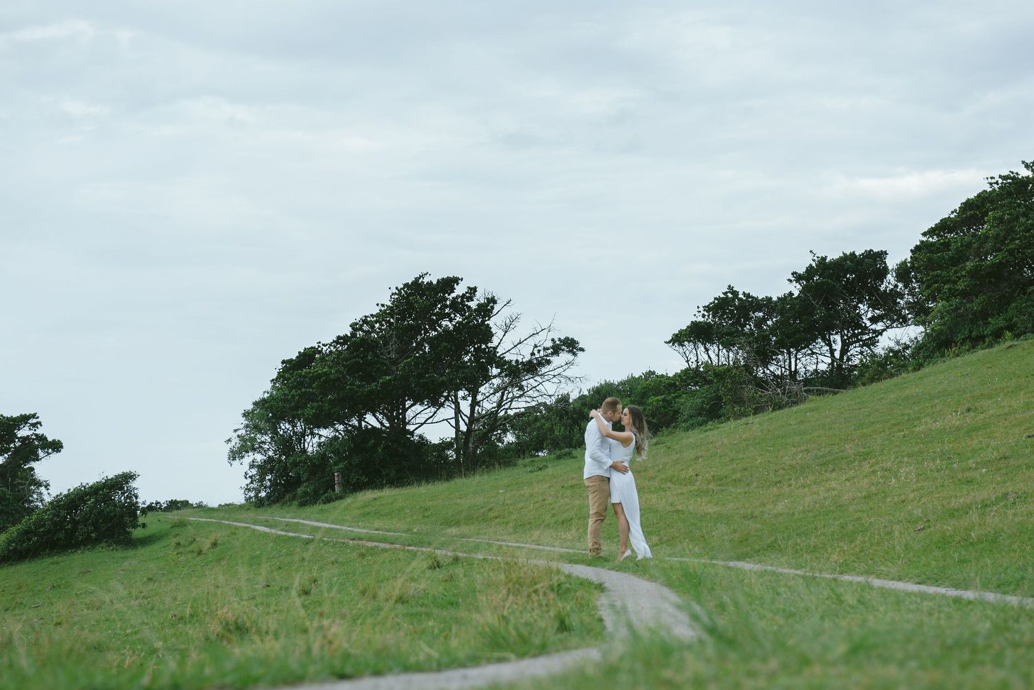 Couple embraces on winding dirt path amid coastal grassland.