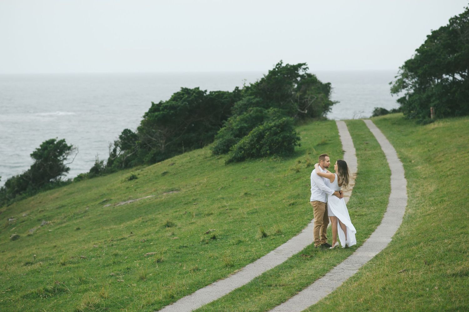 A couple embraces on a winding coastal path overlooking the ocean with green hills and trees in the background.