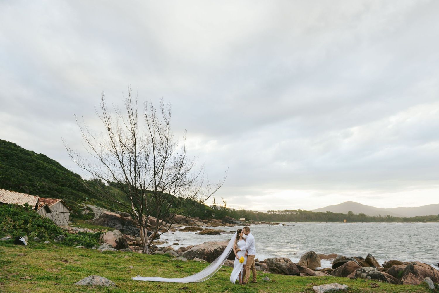 Long flowing white dress catches wind on rocky coastline under cloudy sky.