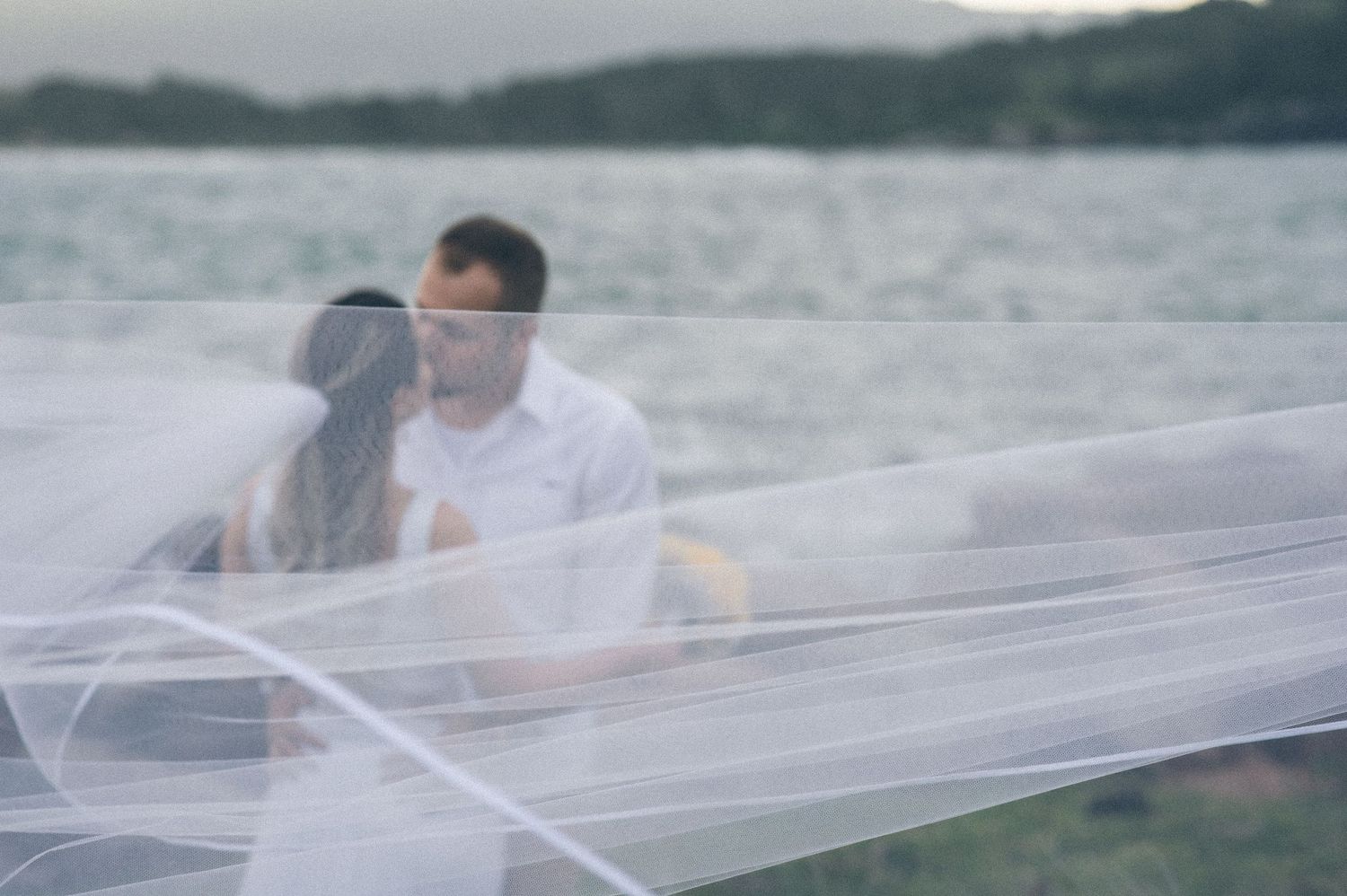 Dreamy portrait through flowing white veil against coastal landscape.