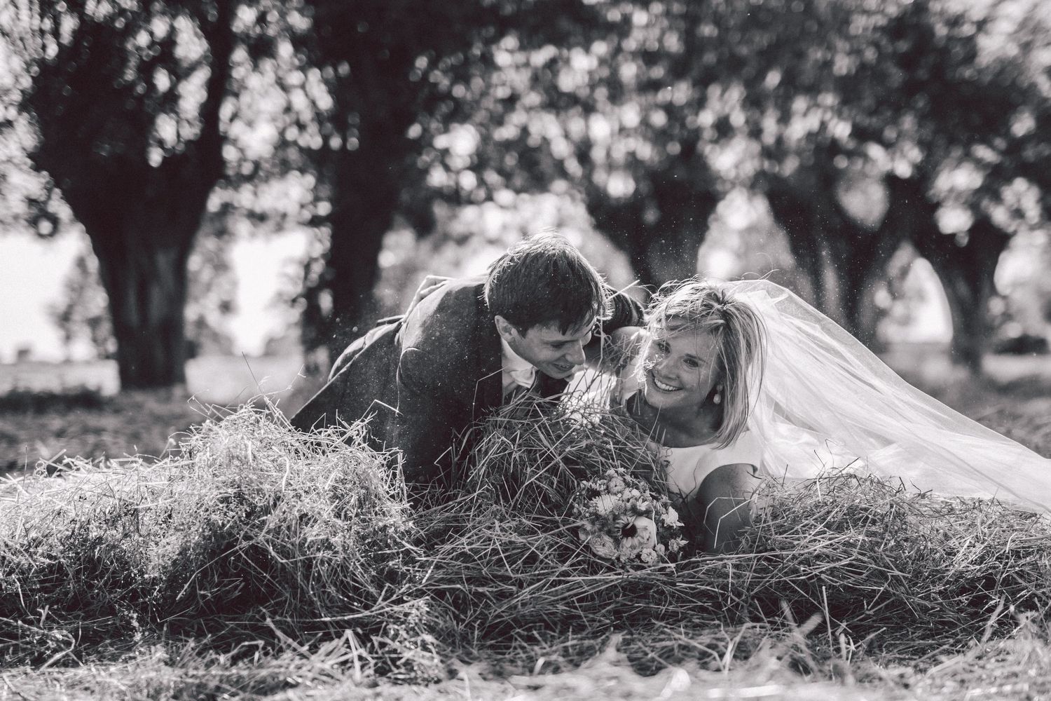 A romantic black and white photo of a couple lying in hay with a wedding veil visible.