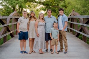 Family of six smiling together on a wooden bridge surrounded by lush green trees during a sunny outdoor photo session.