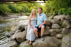 A smiling couple sits on rocks beside a peaceful creek surrounded by lush green trees during golden hour.