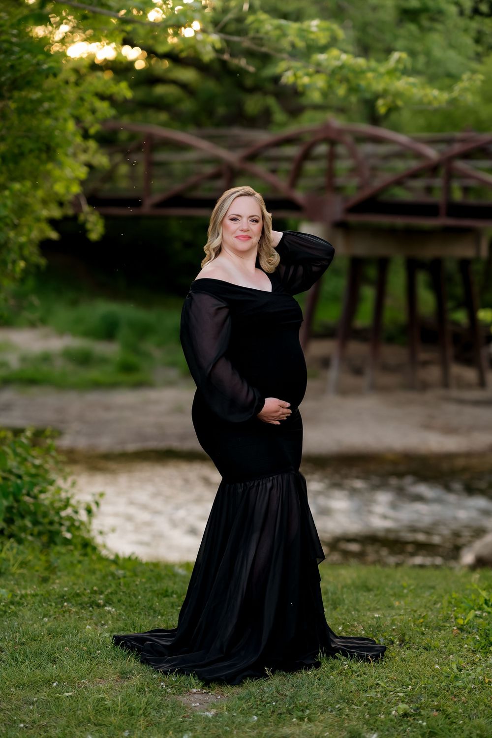 Pregnant woman in elegant black off-shoulder mermaid gown poses outdoors near a bridge surrounded by lush greenery.